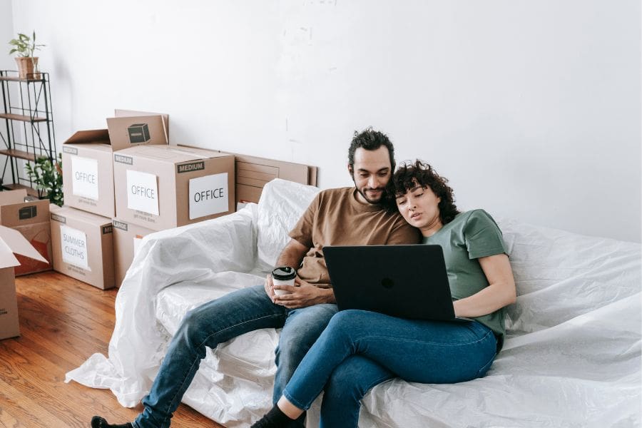 Couple sitting on a couch and browsing on a computer. Moving boxes are in the background.