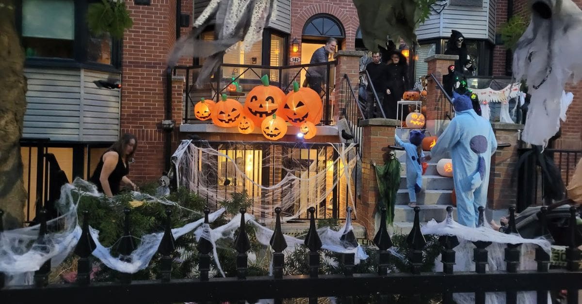 A young child is dressed in a Halloween costume in Canada, trick-or-treating at a decorated home with inflatable pumpkins, spider webs, and ghosts.
