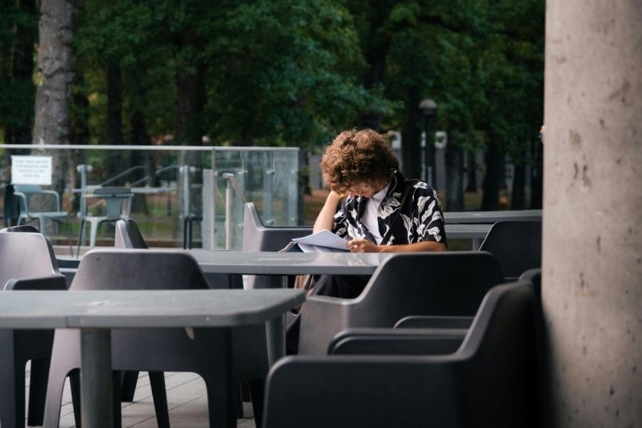 A student sits outdoors on the UBC campus, studying with notebooks and a laptop, surrounded by trees and university buildings.