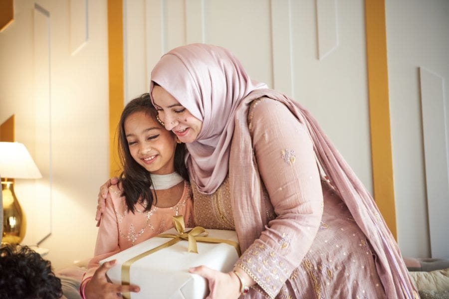 A heartwarming portrait of a Muslim mother and her young daughter smiling together while holding wrapped Eid gifts in a Canadian home.