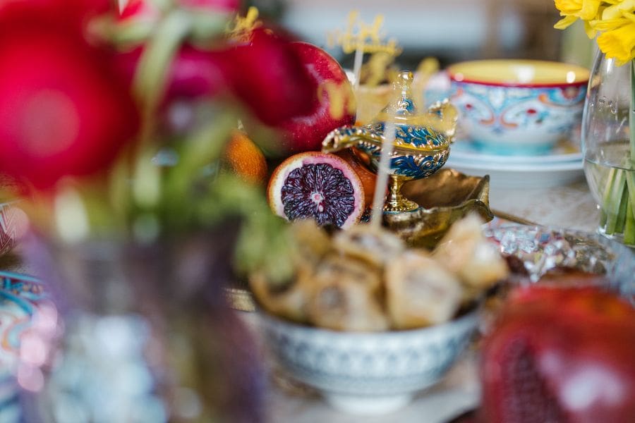 A vibrant close-up of an Eid al-Fitr table featuring a decorative gold lantern, fresh red flowers, blood oranges, and a bowl of traditional sweets like baklava.