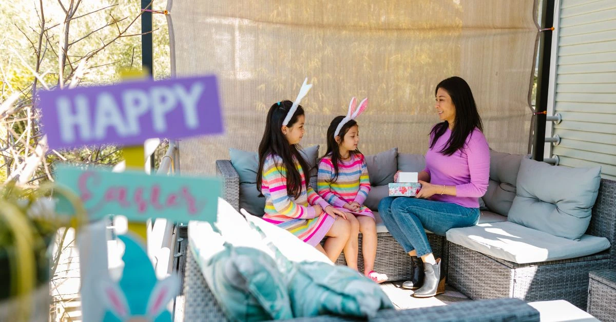 A mother and her two young daughters are wearing bunny ears and sitting on a sunlit patio in Canada. They are exchanging gifts near a Happy Easter sign.