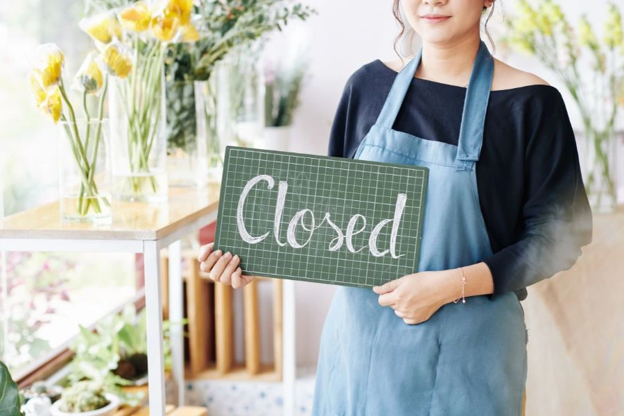 A female shop owner in a blue apron is holding a "Closed" sign in a flower shop, reflecting holiday closures in Canada.