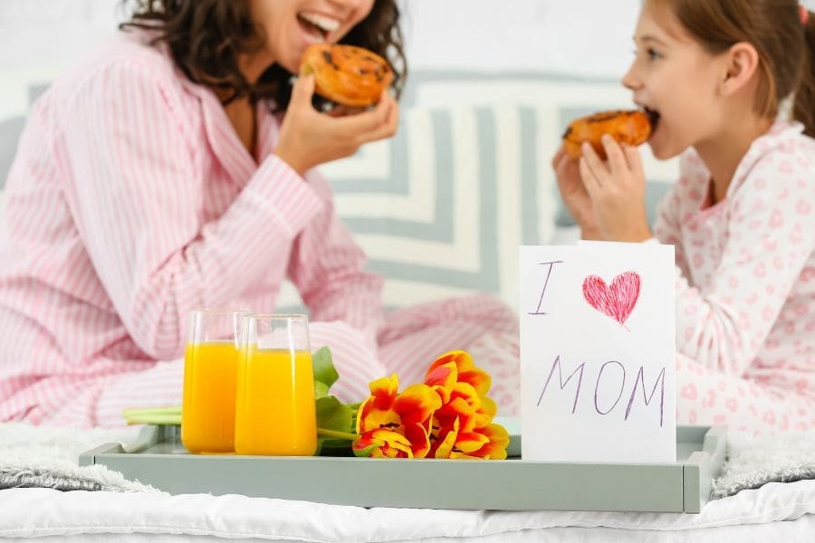 A young girl and her mom are eating breakfast in bed, with a breakfast tray, flowers, and a handmade 'I Heart Mom' card.