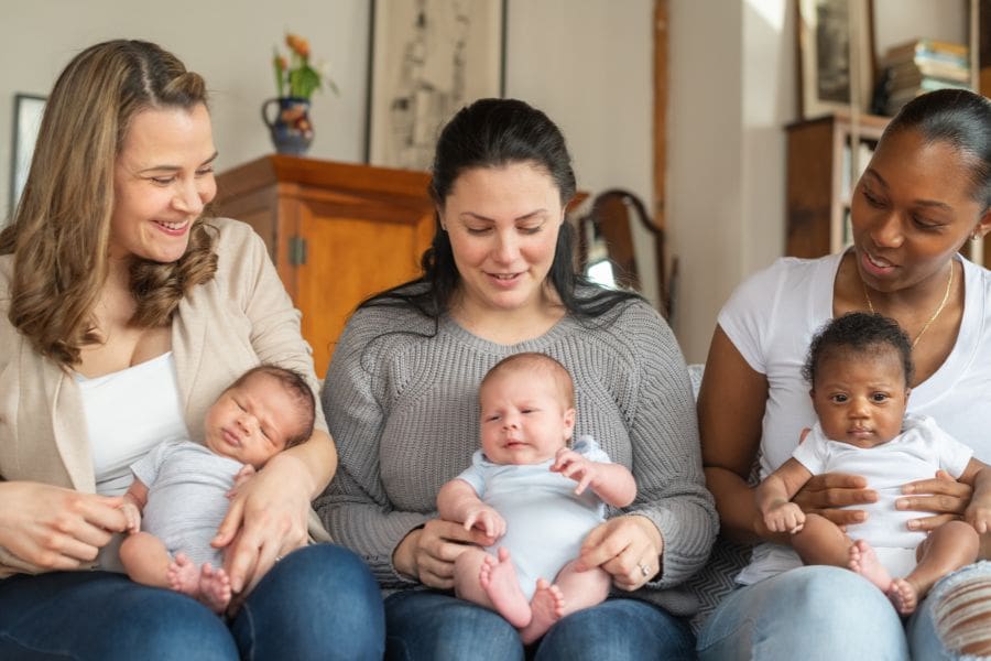 Three diverse mothers sitting together on a couch, each smiling and holding an infant on her lap.