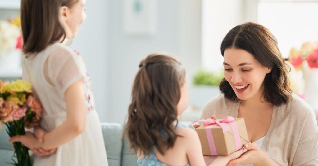 A mother is smiling while her children surprise her with a wrapped gift and flowers on Mother's Day.