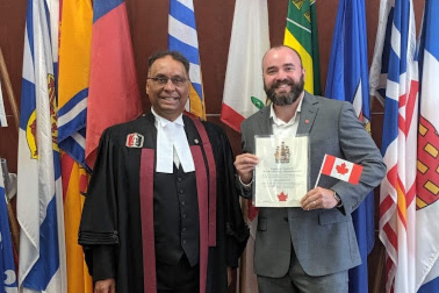 A proud new Canadian citizen holding his citizenship certificate and a small Canadian flag next to a citizenship judge in ceremonial robes.