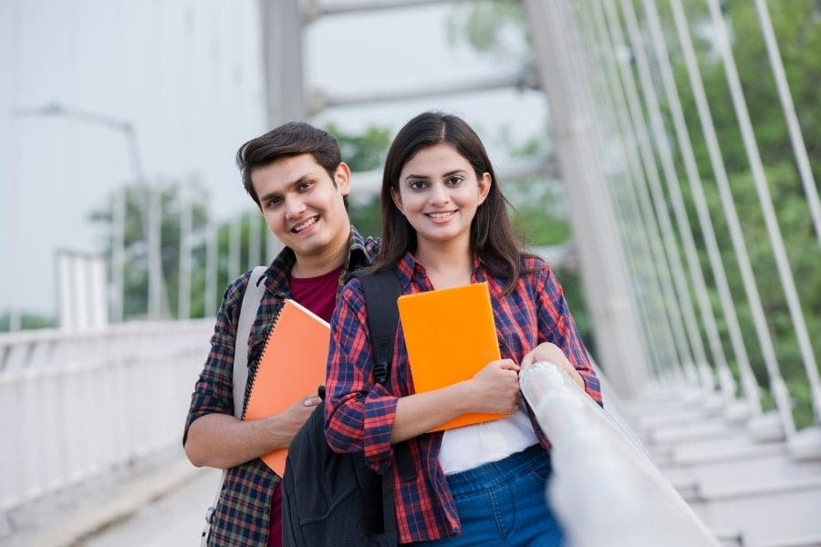Two young international students holding notebooks and smiling on a modern pedestrian bridge in Edmonton.