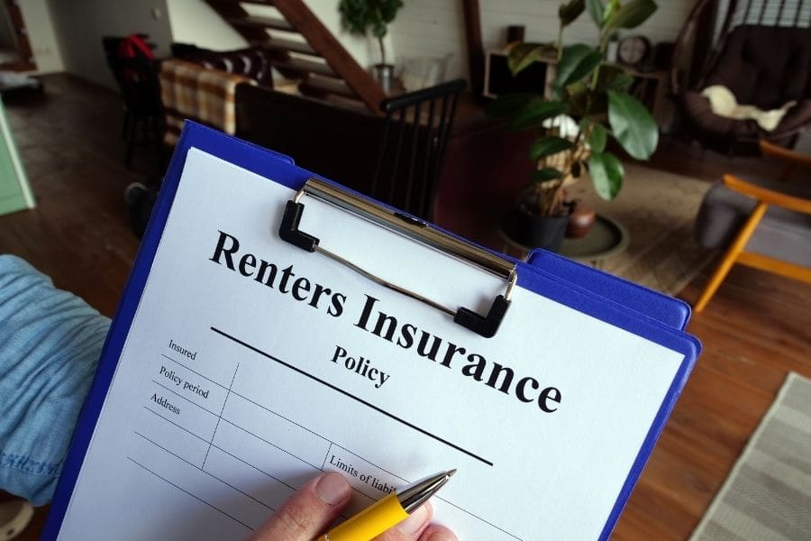 A hand holding a pen over a renters insurance policy document on a clipboard inside a modern apartment living room.