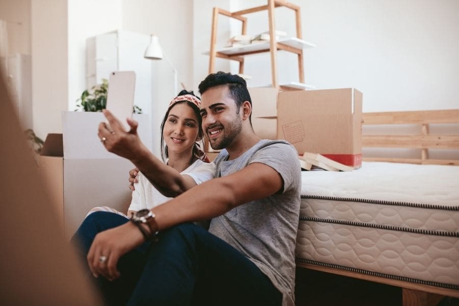 A happy young couple taking a smartphone selfie while sitting on the floor surrounded by moving boxes in their new rental apartment.