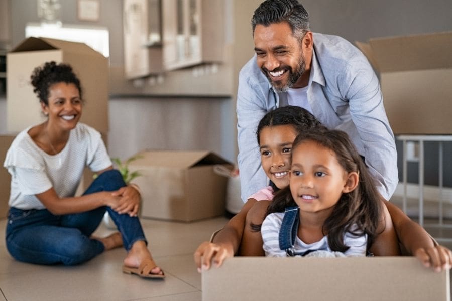 A happy Indian family has moved into a new apartment. The father is pushing his smiling daughters in a moving box, while their mother is watching from aside. 