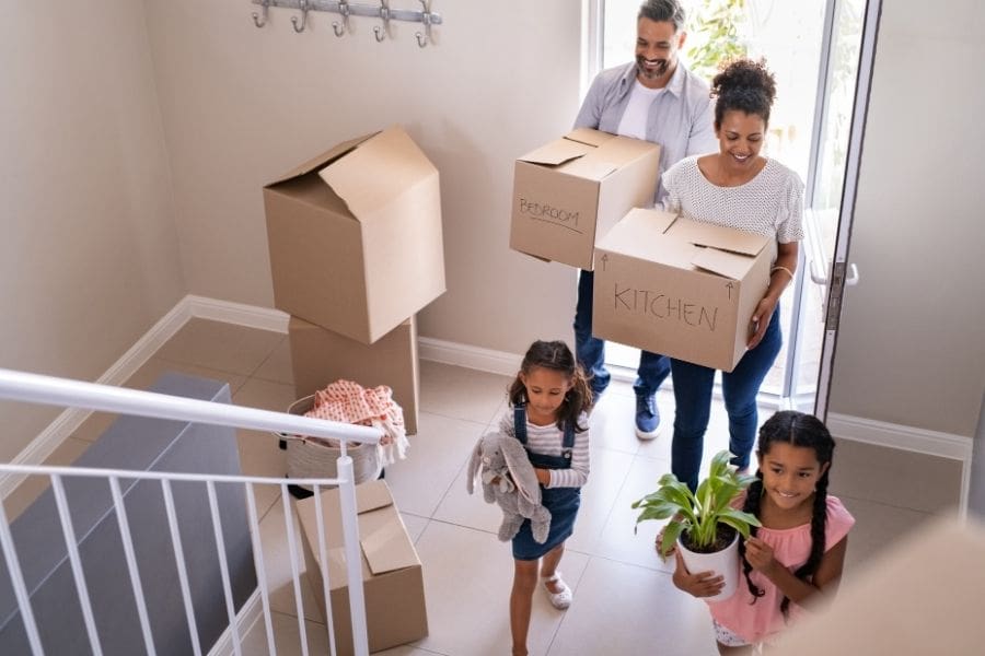 A young family is moving into their new rental home. They parents are carrying moving boxes and their young daughters are carrying a plant and a stuffed animal. 
