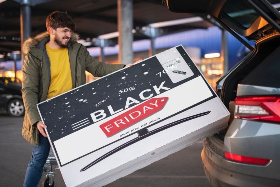 A young man is loading a large TV set into a car trunk after a Black Friday electronics purchase.