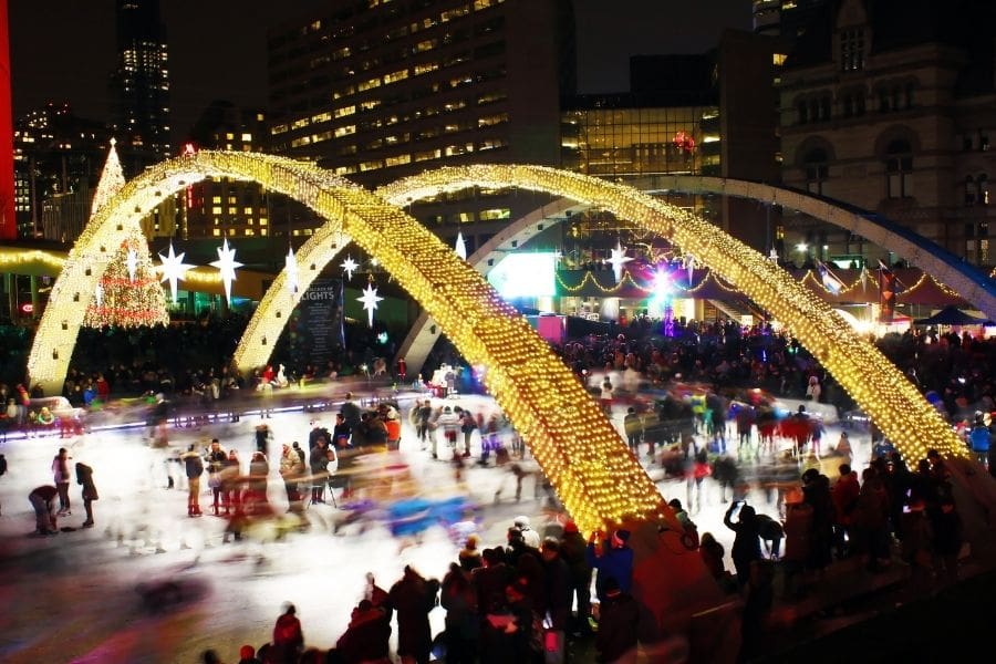People ice skating outdoors at Nathan Phillips Square during the Cavalcade of Lights in Toronto, with Christmas lights and decorations.