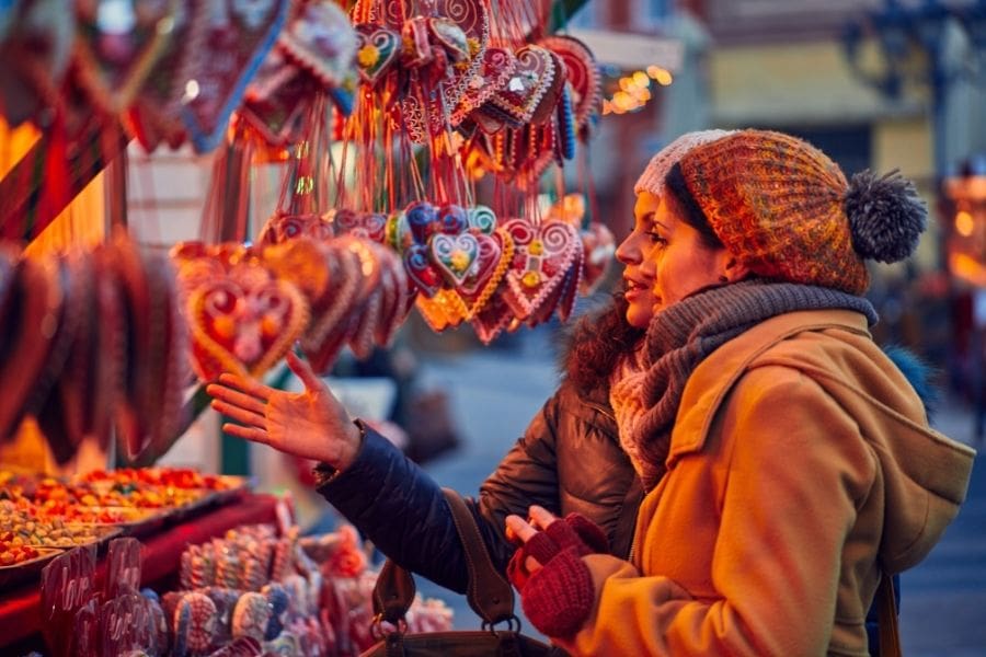 Two newcomers smiling and looking at a colourful candy display at a Christmas market, surrounded by holiday lights and decorations, 2025.