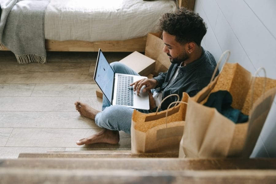 A man sitting on the floor at home, using a laptop to browse the internet, symbolizing budgeting and planning expenses before moving to Canada.