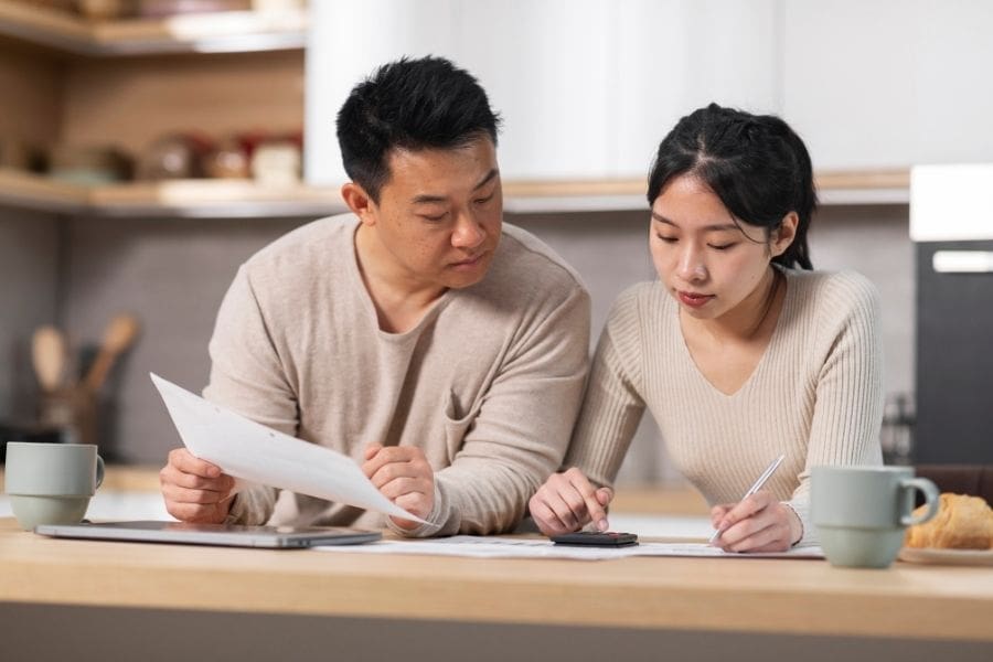 Asian couple calculating monthly rent and living expenses with a calculator, laptop, and coffee mugs while planning their rental budget in Canada