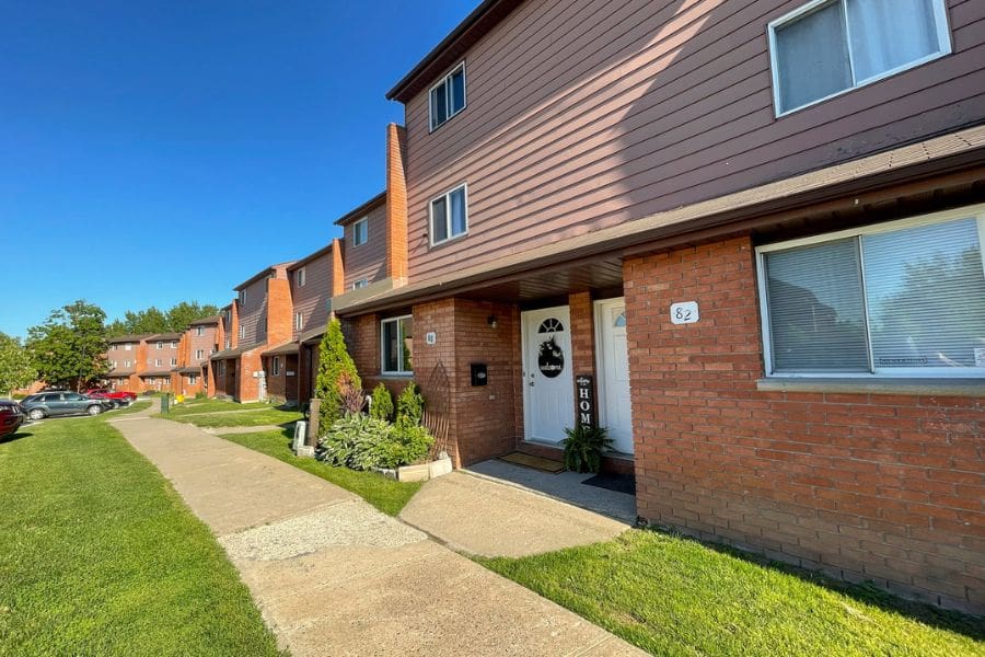 A view of a residential neighbourhood on a bright day, featuring a well-maintained brick house townhouses. Mature trees line the street.