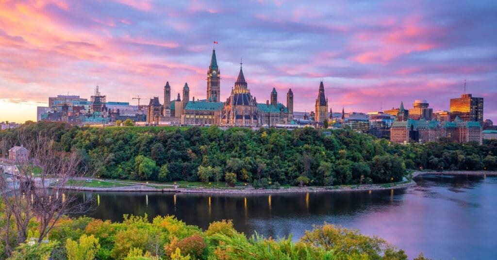 Sunset panorama of Parliament Hill in Ottawa, Ontario, featuring the Peace Tower and the Library of Parliament. The gothic architecture sits above the lush green banks of the Ottawa River, representing the heart of Canada's capital for new arrivals.
