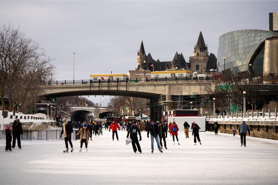 A wide shot of the Rideau Canal Skateway in winter, crowded with people skating on the ice. The Fairmont Château Laurier and Senate of Canada building are visible in the background under a winter sky.