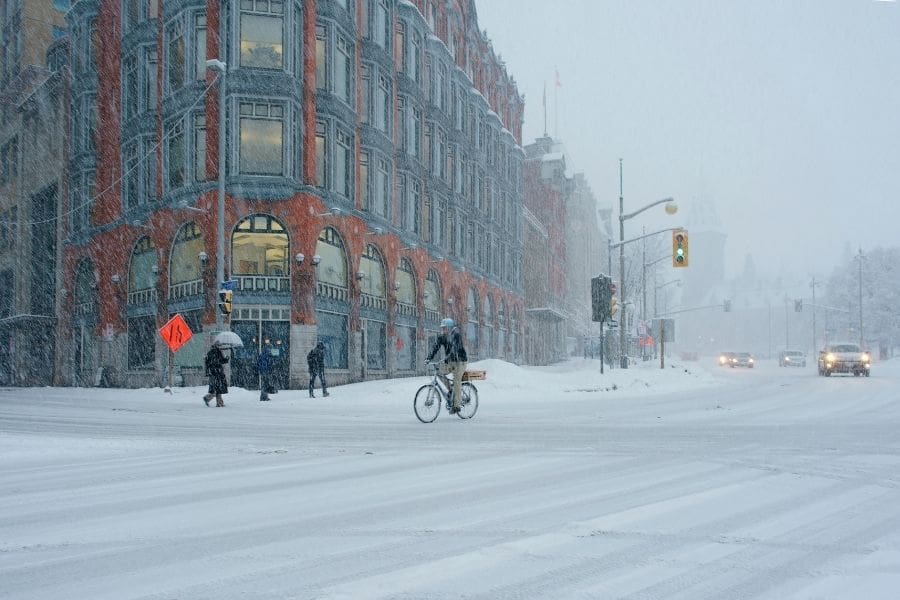 A snowy street scene in downtown Ottawa during a winter storm. A cyclist and pedestrians cross a snow-covered intersection in front of a historic red brick building, showing the reality of winter life in Canada's capital.
