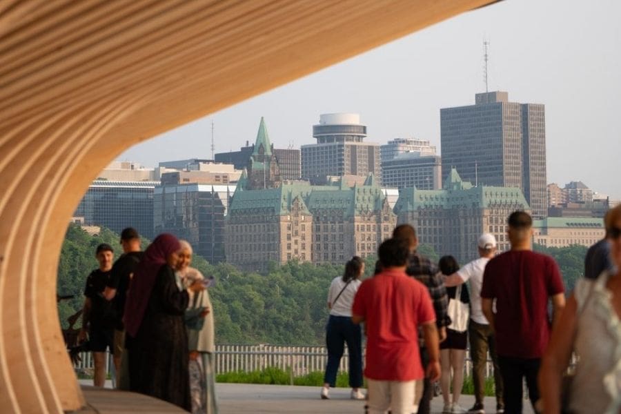 A group of diverse people, including some in traditional Islamic dress, stand at a modern wooden lookout point. In the background, the green-roofed Confederation Building and the downtown Ottawa skyline are visible under a soft evening light.