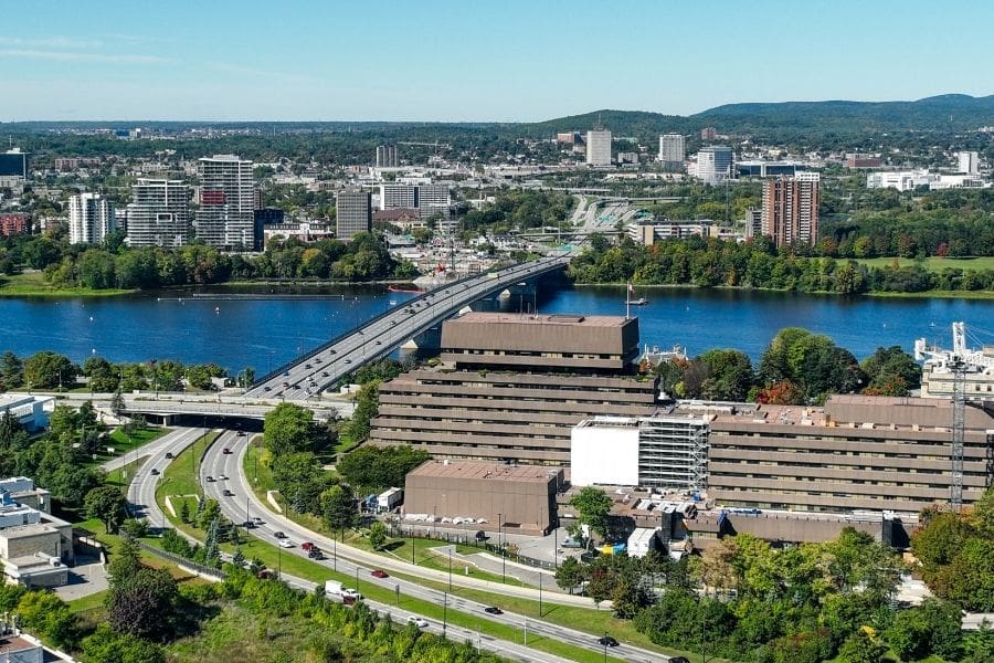 Aerial view of the Macdonald-Cartier Bridge spanning the Ottawa River between Ontario and Quebec. The image shows multiple lanes of traffic, urban buildings in the foreground, and the Gatineau Park hills in the distance. Ottawa rent prices are higher than Gatineau, but newcomers should be aware of the trade-offs.