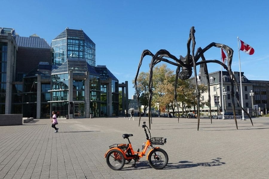 A giant bronze spider sculpture titled 'Maman' stands on a stone plaza in front of the glass towers of the National Gallery of Canada. An orange electric tricycle is parked in the foreground under a clear blue sky.