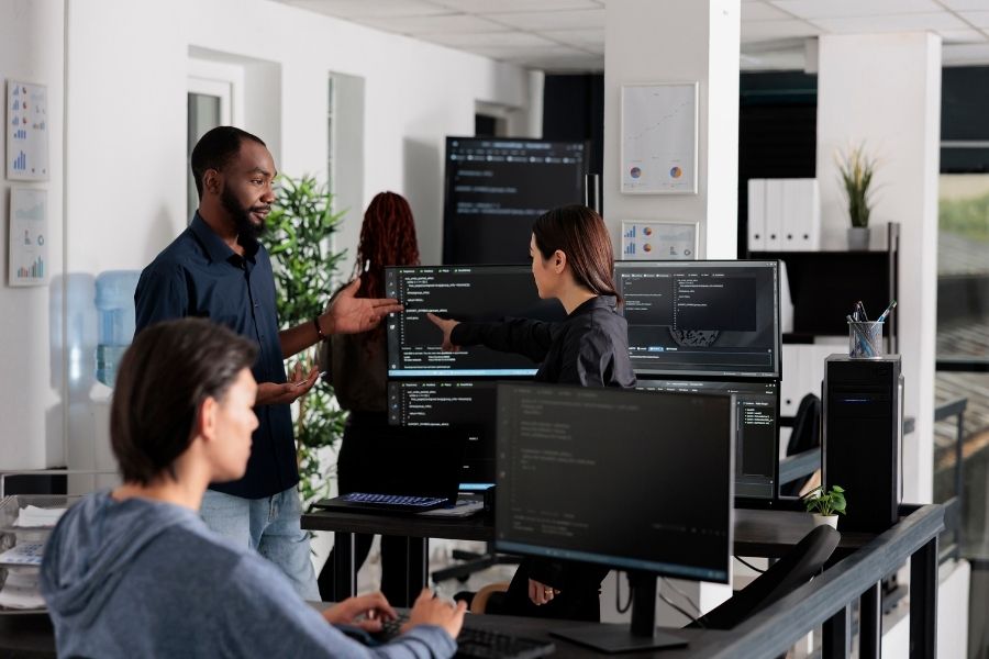 A diverse team of software developers in a modern office discussing code on multiple computer monitors
