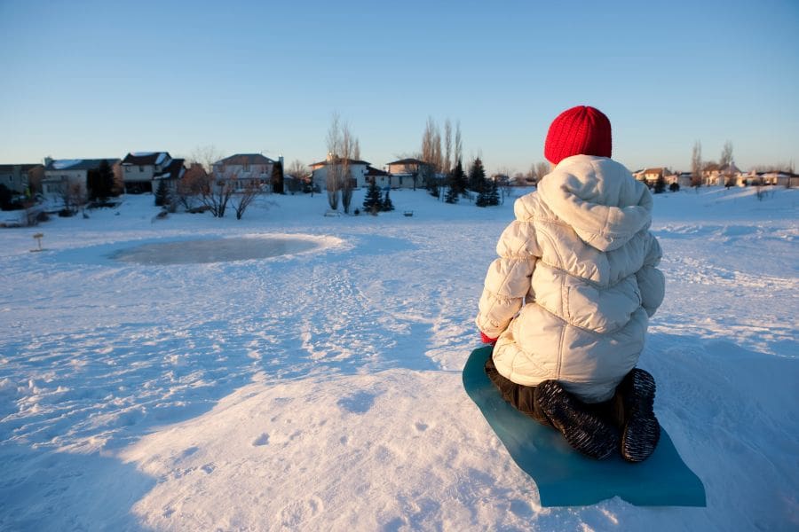 A child in a white winter coat and red hat sitting on a blue sled in the snow. A residential neighbourhood is in the background.