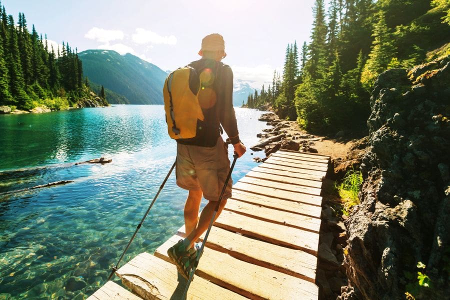 A young man is hiking with a yellow backpack and poles near a turquoise Canadian mountain lake.