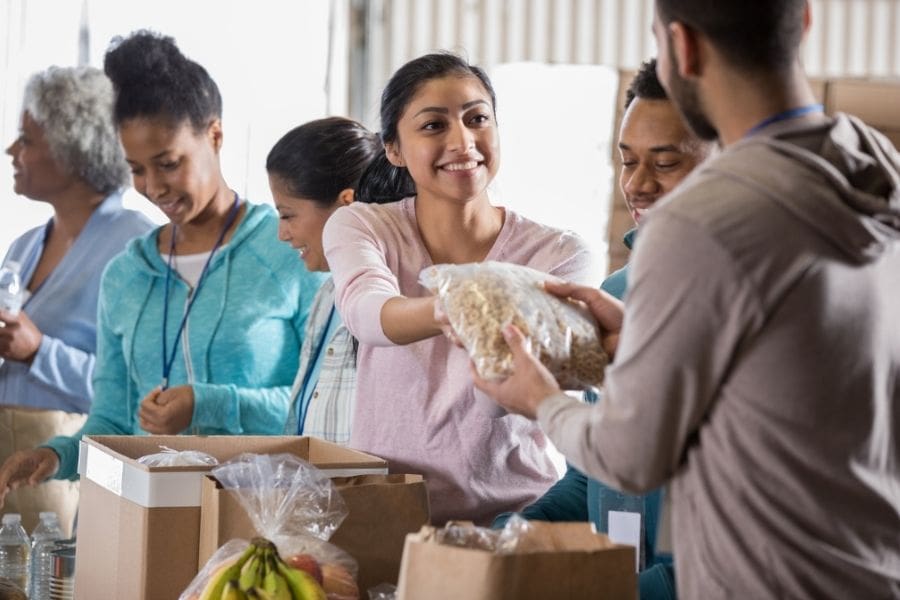 A smiling young woman handing a food donation bag to a person, surrounded by other diverse volunteers.