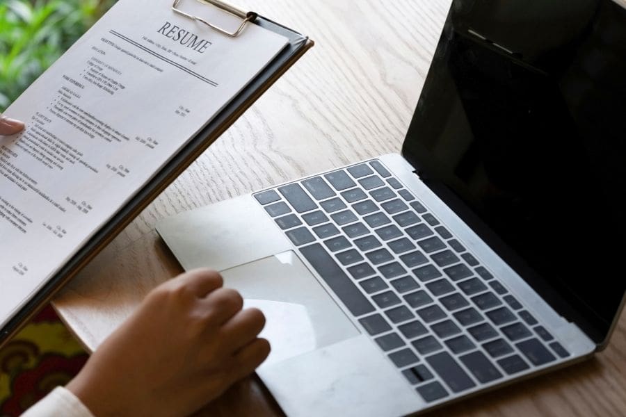 A person holding a resume on a clipboard next to a laptop, preparing a job application.