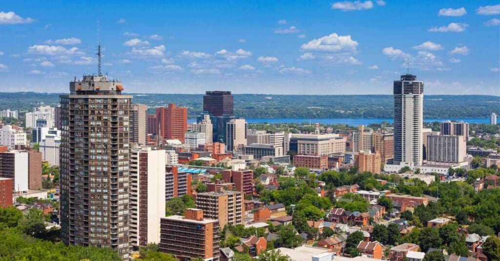 A panoramic high-angle view of the Hamilton, Ontario skyline. It shows Landmark Place and other downtown high-rises, surrounded by green space with Lake Ontario in the distant background under a blue sky.