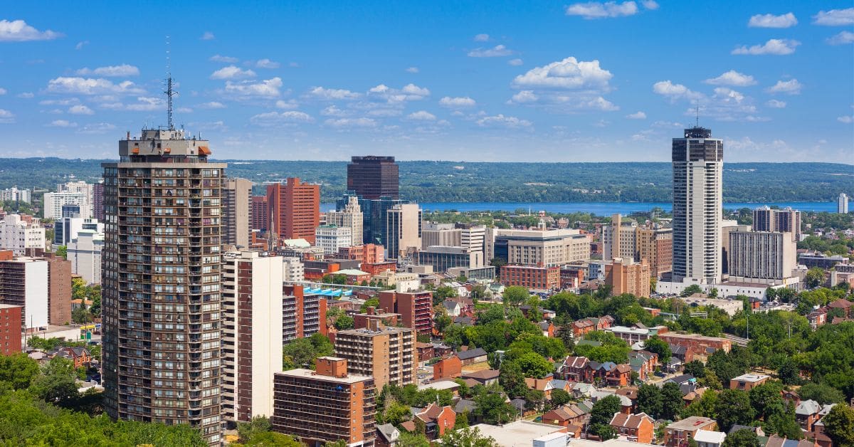 A panoramic high-angle view of the Hamilton, Ontario skyline. It shows Landmark Place and other downtown high-rises, surrounded by green space with Lake Ontario in the distant background under a blue sky.
