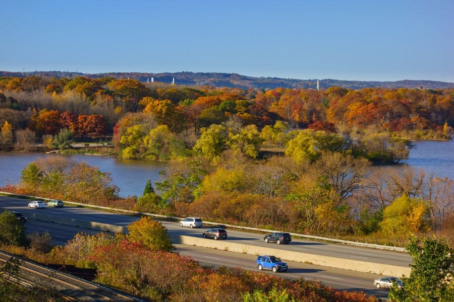 A high-angle view of Highway 403 in Hamilton, Ontario. Cars are driving near the scenic Cootes Paradise and a calm body of water with scenic and colourful fall foliage. 