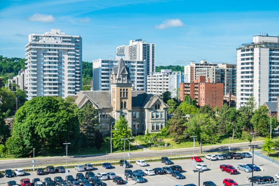 Central Public School in downtown Hamilton. A stone Victorian building with a clock tower, surrounded by green trees and modern apartment buildings under a blue sky.