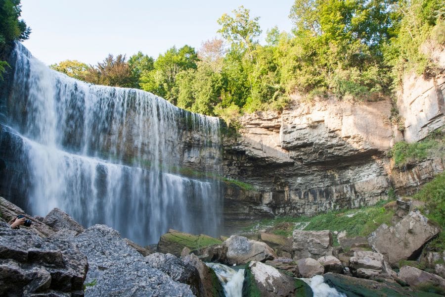 A large, wide tiered waterfall cascading over a rocky limestone cliff into a river basin. There are surrounding lush green trees and grey boulders under a bright sky in Ontario.