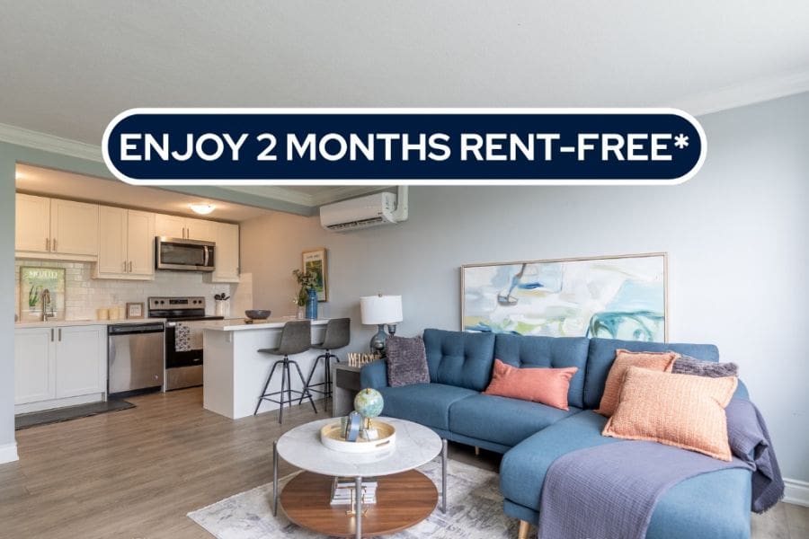 A modern open-concept apartment living room and kitchen in Hamilton, Ontario. It shows a blue sofa and white cabinetry with a banner reading 'Enjoy 2 Months Rent-Free.'