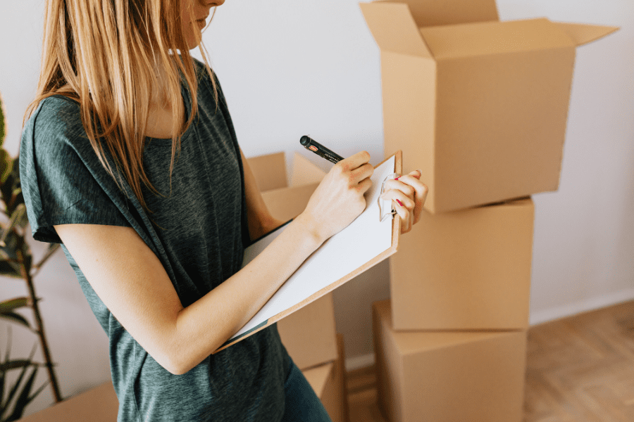 Person making a checklist beside moving boxes before moving to Canada. The BSF186 is also known as the Settler's Effect List.