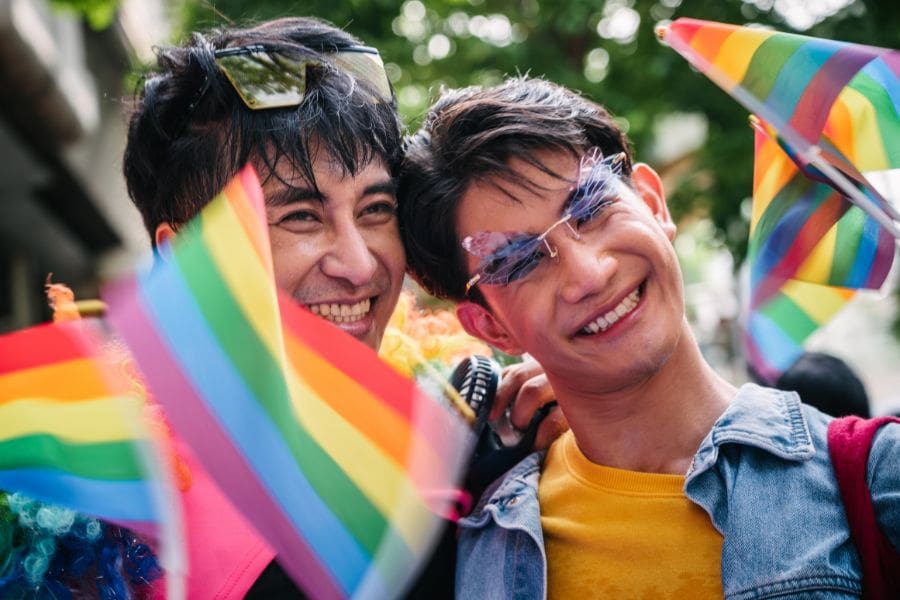 Two joyful East Asian men smiling together and waving rainbow flags at an outdoor Pride parade festival.