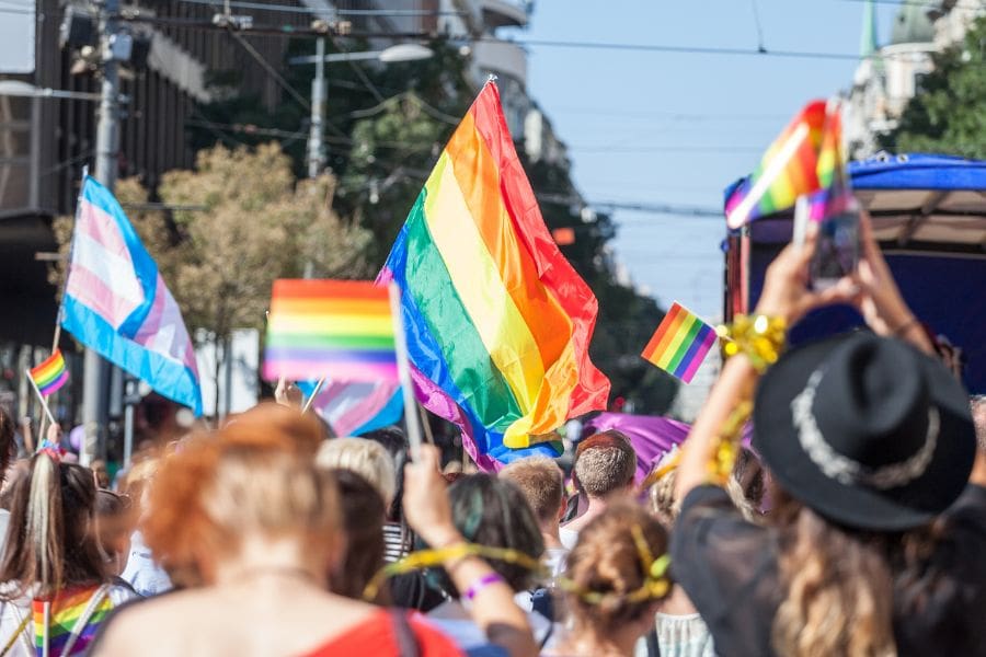 A crowded city street during a Canada Pride parade with people waving rainbow flags and a transgender pride flag under a bright blue sky.
