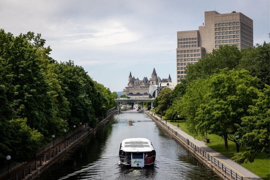 An electric passenger tour boat navigating down the calm waters of the Rideau Canal in Ottawa, with lush green tree-lined pathways on both sides and the historic Château Laurier hotel visible in the background under an overcast sky.