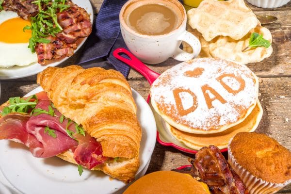 A festive Father's Day breakfast spread featuring pancakes with powdered sugar that reads "I love DAD," waffles, and a croissant sandwich.