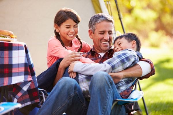 A happy father laughing and hugging his son and daughter outside their tent while camping on Father's Day in Canada.