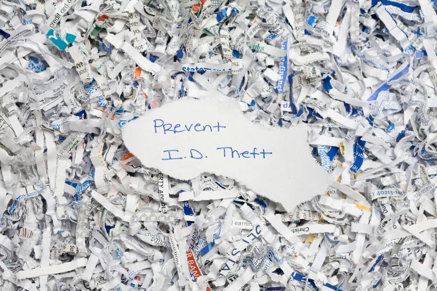 Hand-torn pieces of paper with the handwritten words Prevent I.D. Theft resting on top of a dense background of shredded white and blue documents.