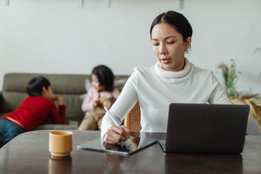 A female newcomer sitting at a desk with a laptop, using a stylus to fill out a Canadian rental application guide and social insurance number application online while her two children sit on a couch in the background.