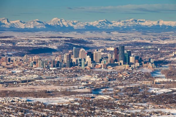 A high-angle winter view of the downtown Calgary skyline and city skyscrapers under a blue sky, with the snow-covered Rocky Mountains in the distant background. Perfect for newcomers exploring options for living in Calgary.