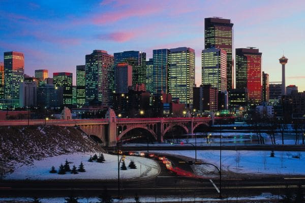 The downtown city skyline illuminated at twilight during winter, featuring the Centre Street Bridge and the Calgary Tower under a colorful dusk sky.