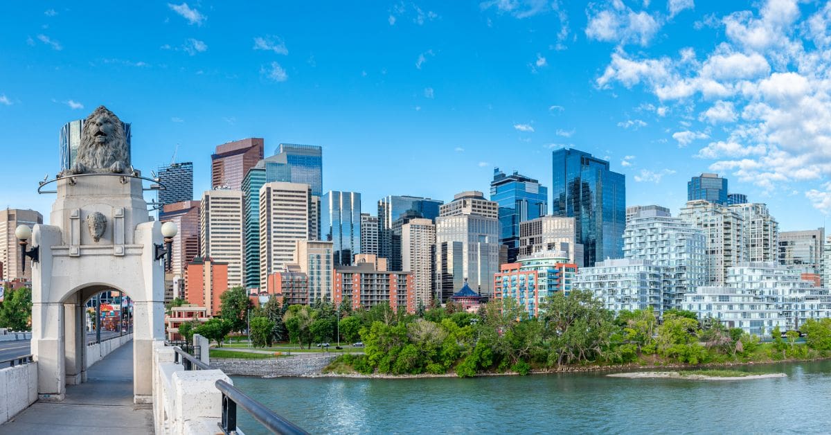 A wide view of the downtown Calgary skyline from the Centre Street Bridge, showing skyscrapers, green trees, and the Bow River under a bright blue sky. An excellent overview for anyone planning on living in Calgary.